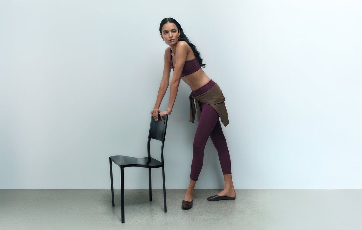 Model leaning on a chair in a fig organic stretch sports bra and leggings – MATE the Label
