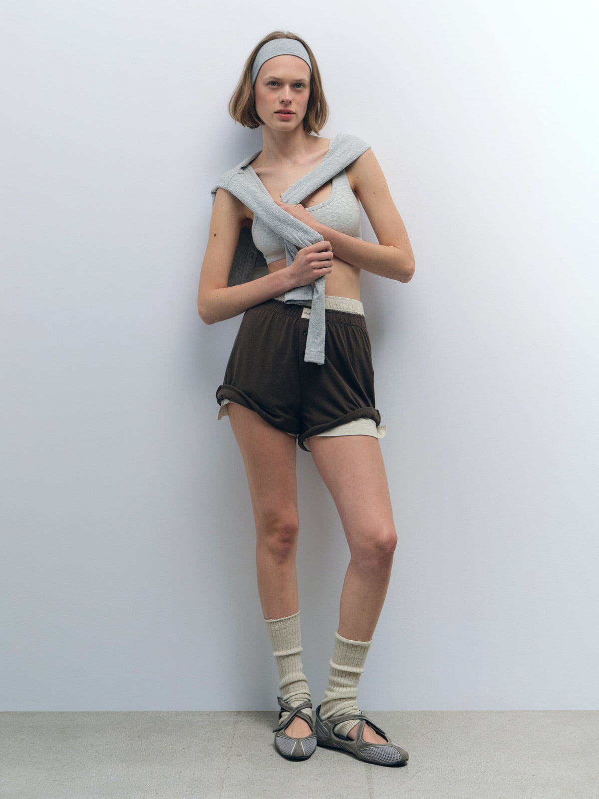 Woman in a light grey Organic Comfort Scoop Bralette, dark brown shorts, and a matching grey sweater.