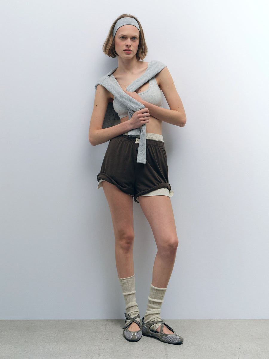 Woman in a light grey Organic Comfort Scoop Bralette, dark brown shorts, and a matching grey sweater.