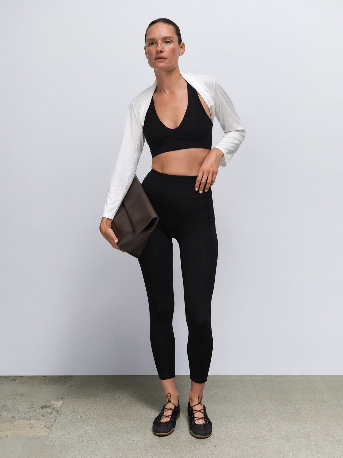 A woman stands against a light gray studio wall wearing a black sports bra and black leggings, with a white cropped cardigan draped off her shoulders and black lace-up flats. She holds a dark brown clutch.