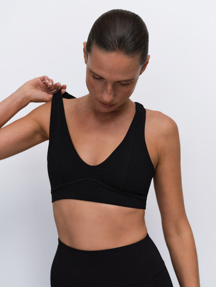 A woman wearing a black sports bra and matching black bottoms, shown from the chest up against a plain light gray studio background. She is adjusting the shoulder strap of the sports bra.