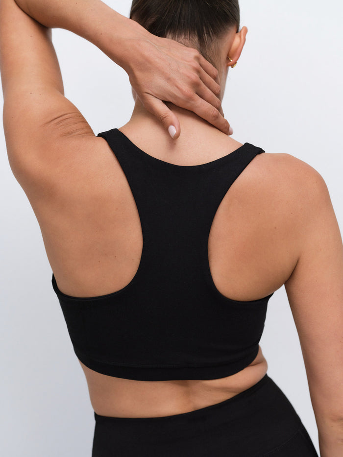 Rear view of a woman wearing a black racerback crop top and black bottoms, with one arm raised over her head, against a light gray studio background.