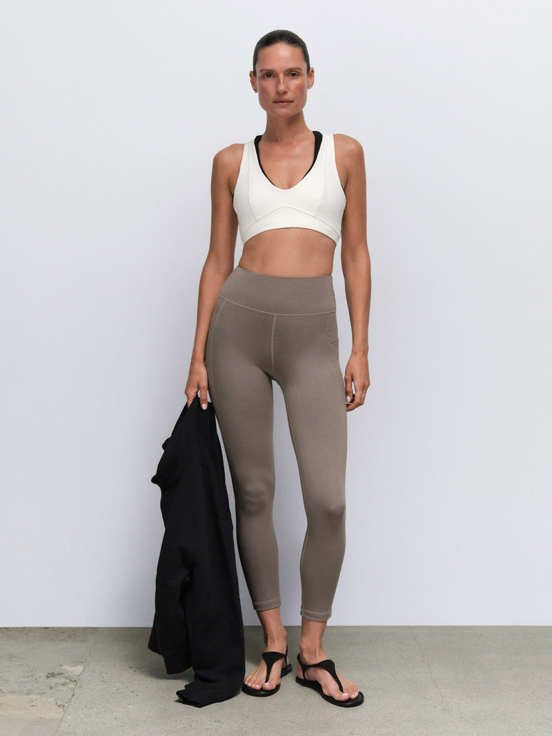 A woman stands against a light gray wall wearing a white sports bra with black trim and taupe high-waisted leggings, holding a black cardigan. She is wearing black flip-flop sandals.