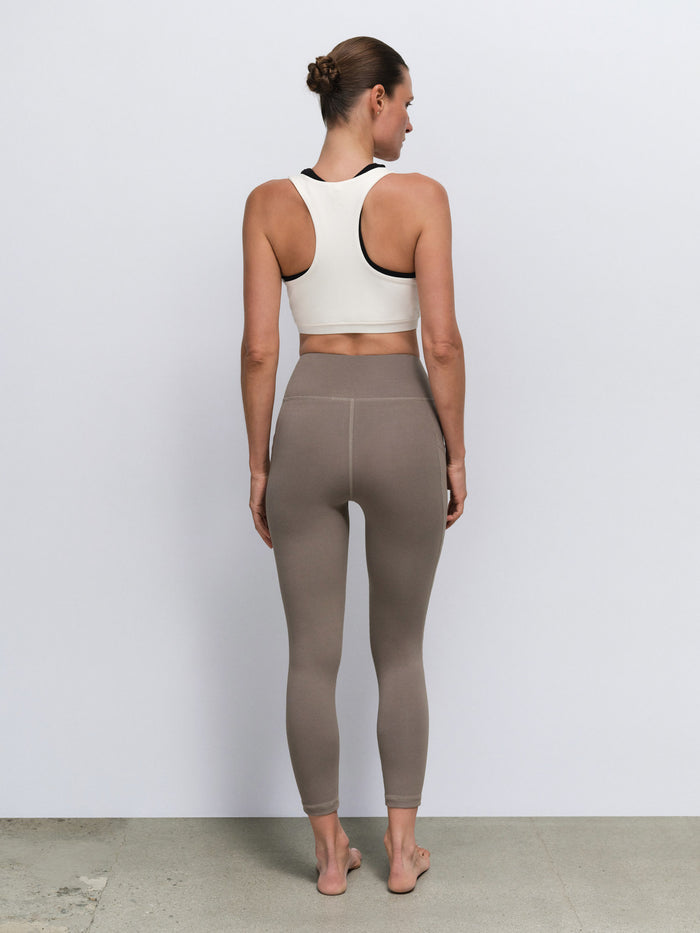 A woman viewed from behind wearing a white sports bra with black trim and high-waisted taupe leggings, standing against a plain light gray wall.