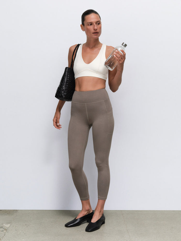 A woman modeling high-waisted taupe gray leggings, paired with a white sports bra, black flats, a black crocodile-embossed shoulder bag, and holding a clear water bottle against a light gray studio background.