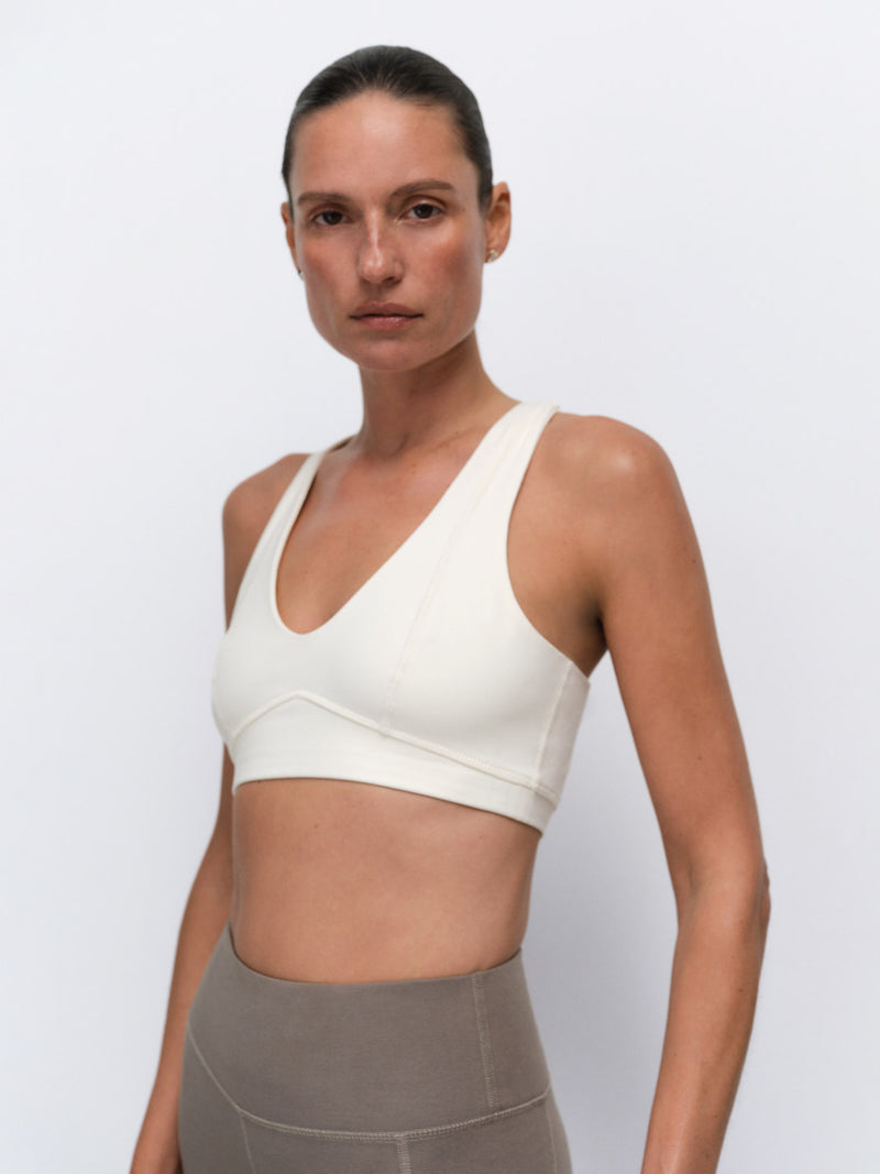 A woman modeling a cream-colored sports bra against a light gray studio background.