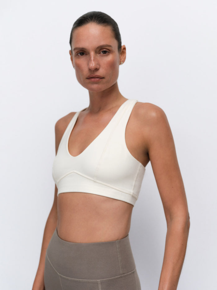 A woman modeling a cream-colored sports bra against a light gray studio background.