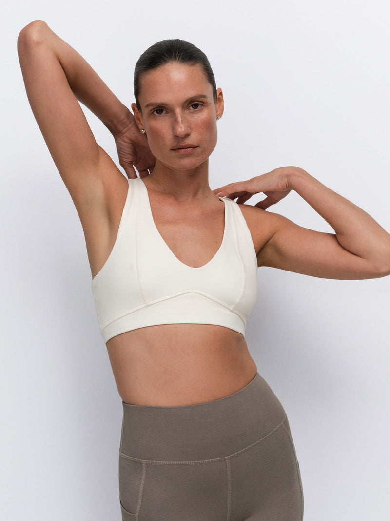 A woman modeling a cream-colored sports bra and taupe-gray high-waisted leggings against a light gray studio background.