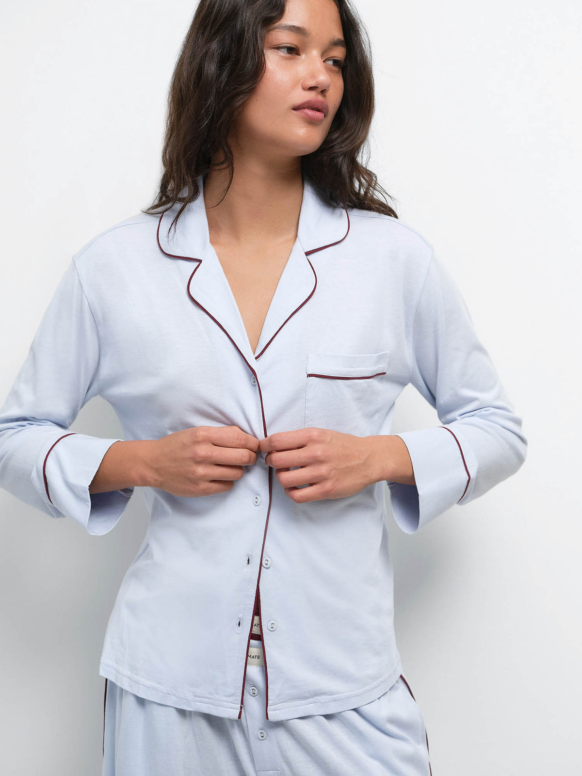 Model buttoning the top of the Tencel Sleep Set in Powder with Garnet piping