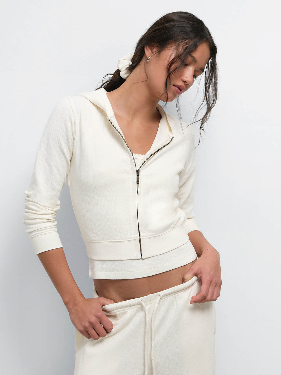 Model looking down with a hand in her pocket, wearing the Organic Pointelle Shrunken Hoodie and Pant in Cream