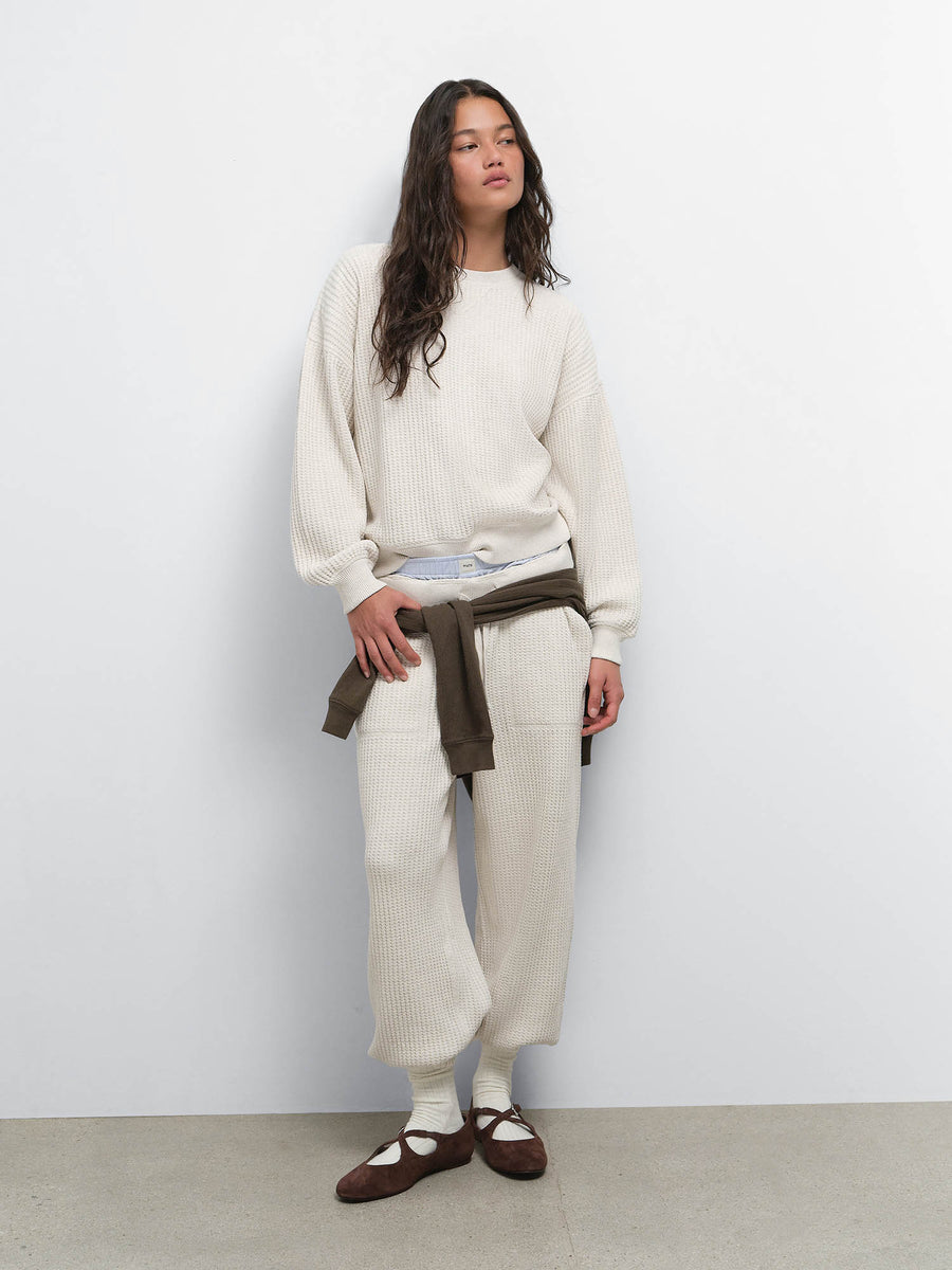 Model with a brown sweater tied at her waist, wearing the Organic Waffle Crewneck Sweatshirt and Sweatpant in Oat