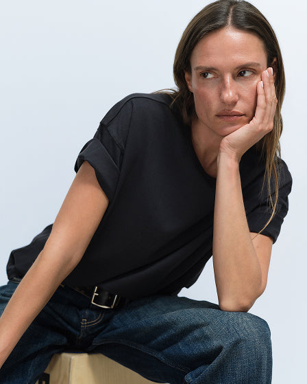 Model wearing a black t-shirt and jeans seated with chin resting on hand
