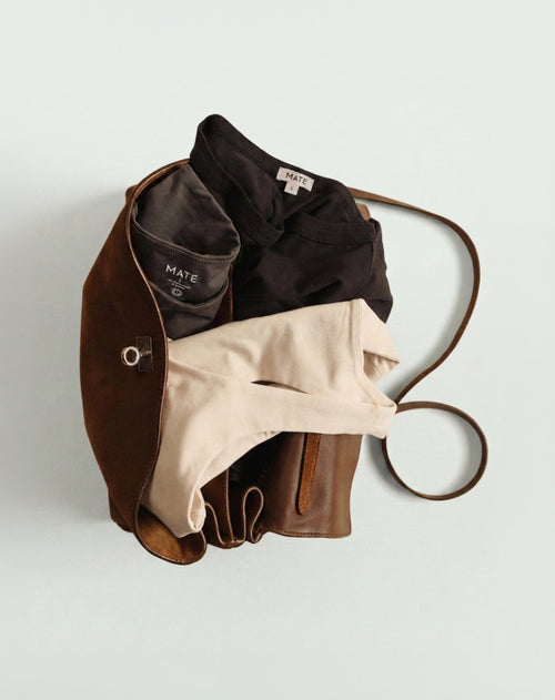 MATE flat lay of a brown vegan leather tote bag with a black cropped top and cream trousers, on a sage green background
