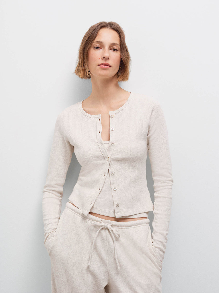 Woman wearing cream pointelle cardigan and matching pants, hands in pockets, front view in studio