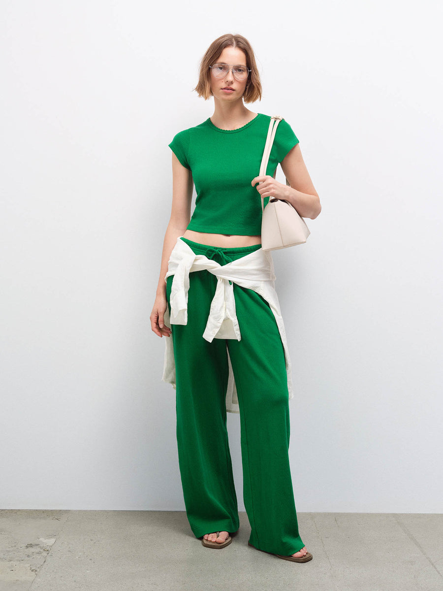 Full-length front view of a woman wearing a green pointelle top and wide-leg pants with glasses and a cream bag