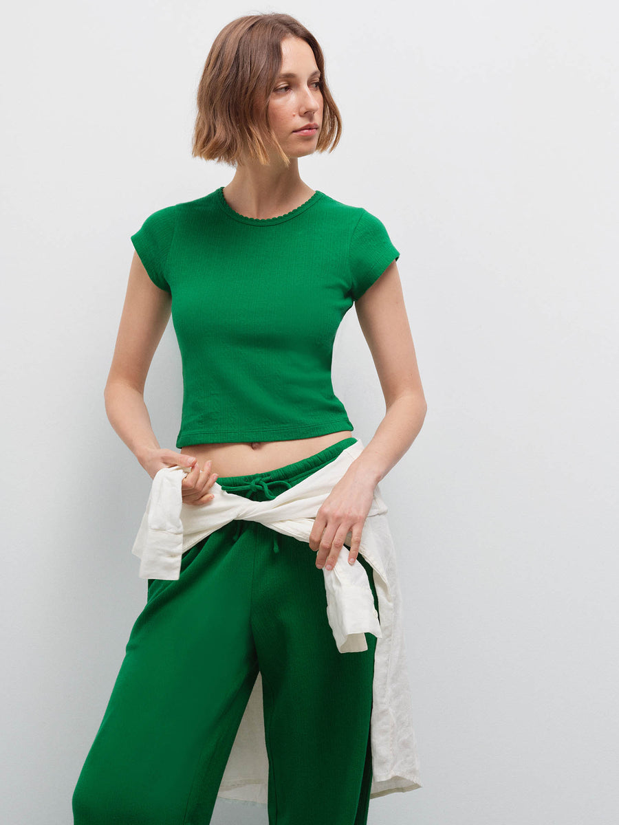 Woman wearing a green pointelle cap-sleeve top and matching wide-leg pants, holding a cream shirt tied at the waist