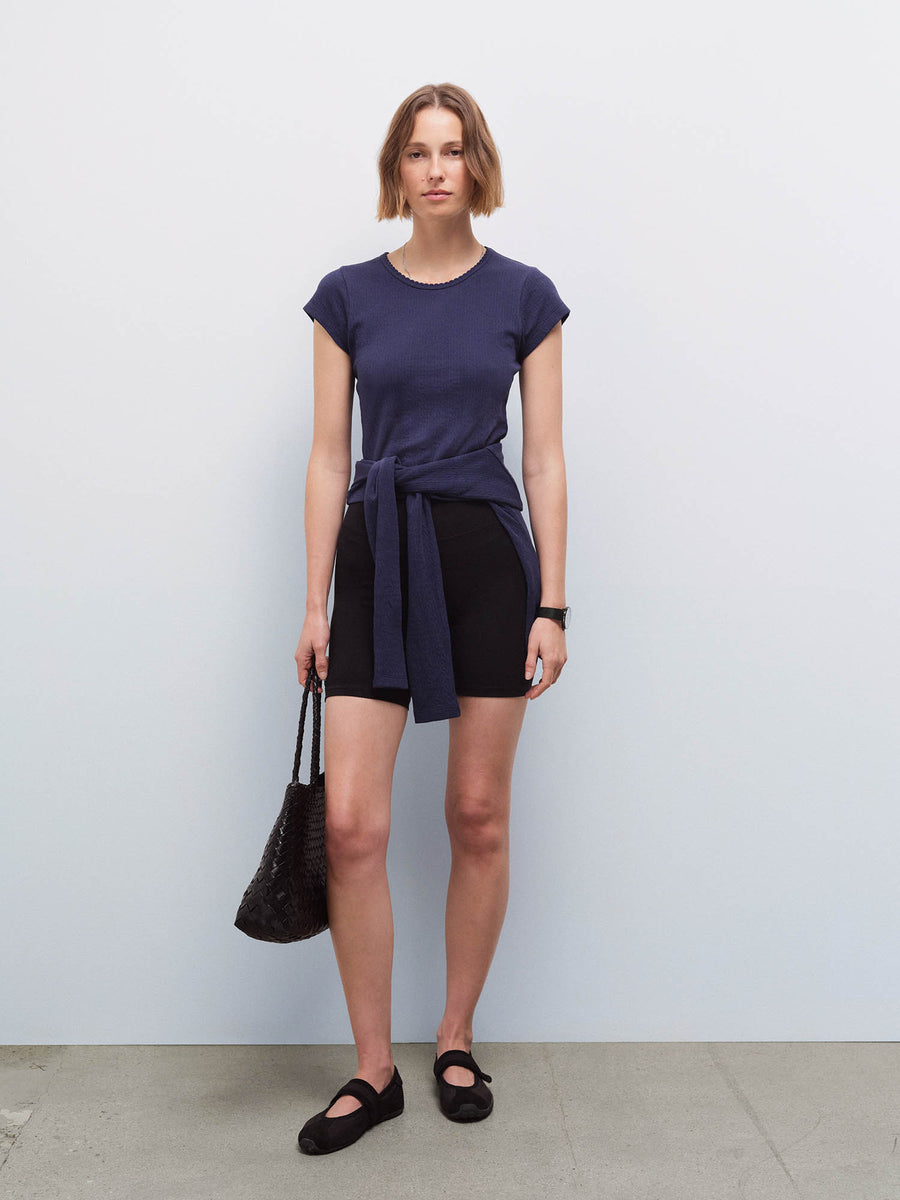 Full-length front view of a woman wearing a navy pointelle top tied at the waist over black shorts, carrying a woven bag