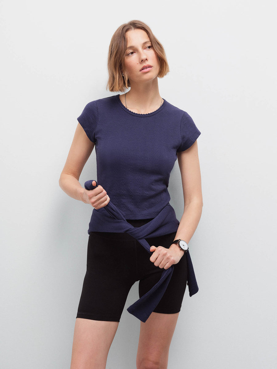 Woman wearing a navy pointelle top tied at the waist over black biker shorts, holding the tie ends