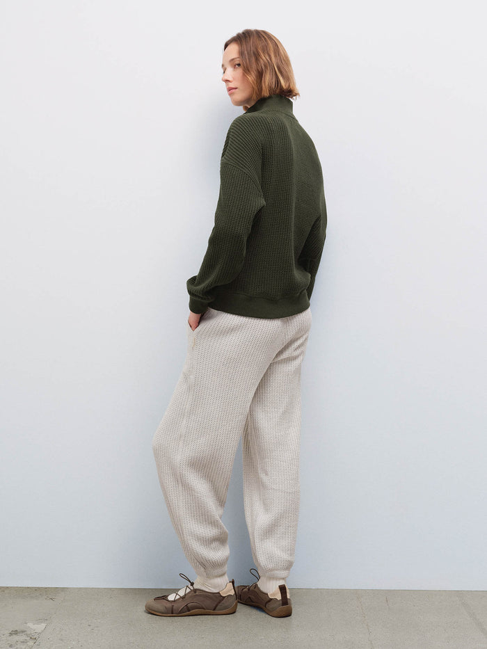 Woman wearing cream waffle-knit sweatpants and olive half-zip sweater, back view in studio