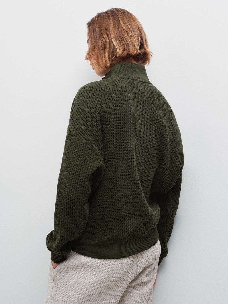 Woman wearing olive waffle-knit half-zip sweatshirt, back view showing collar and waffle texture in studio