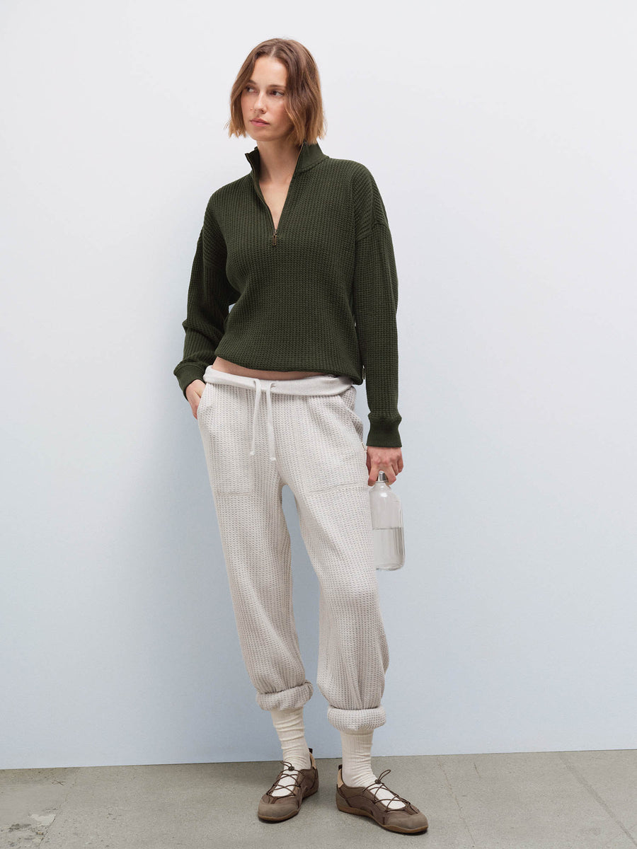 Woman wearing cream waffle-knit sweatpants with olive half-zip sweater, holding a water bottle, front view in studio