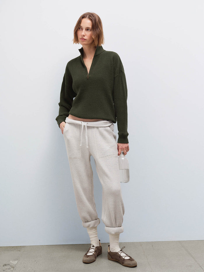Woman wearing cream waffle-knit sweatpants with olive half-zip sweater, holding a water bottle, front view in studio