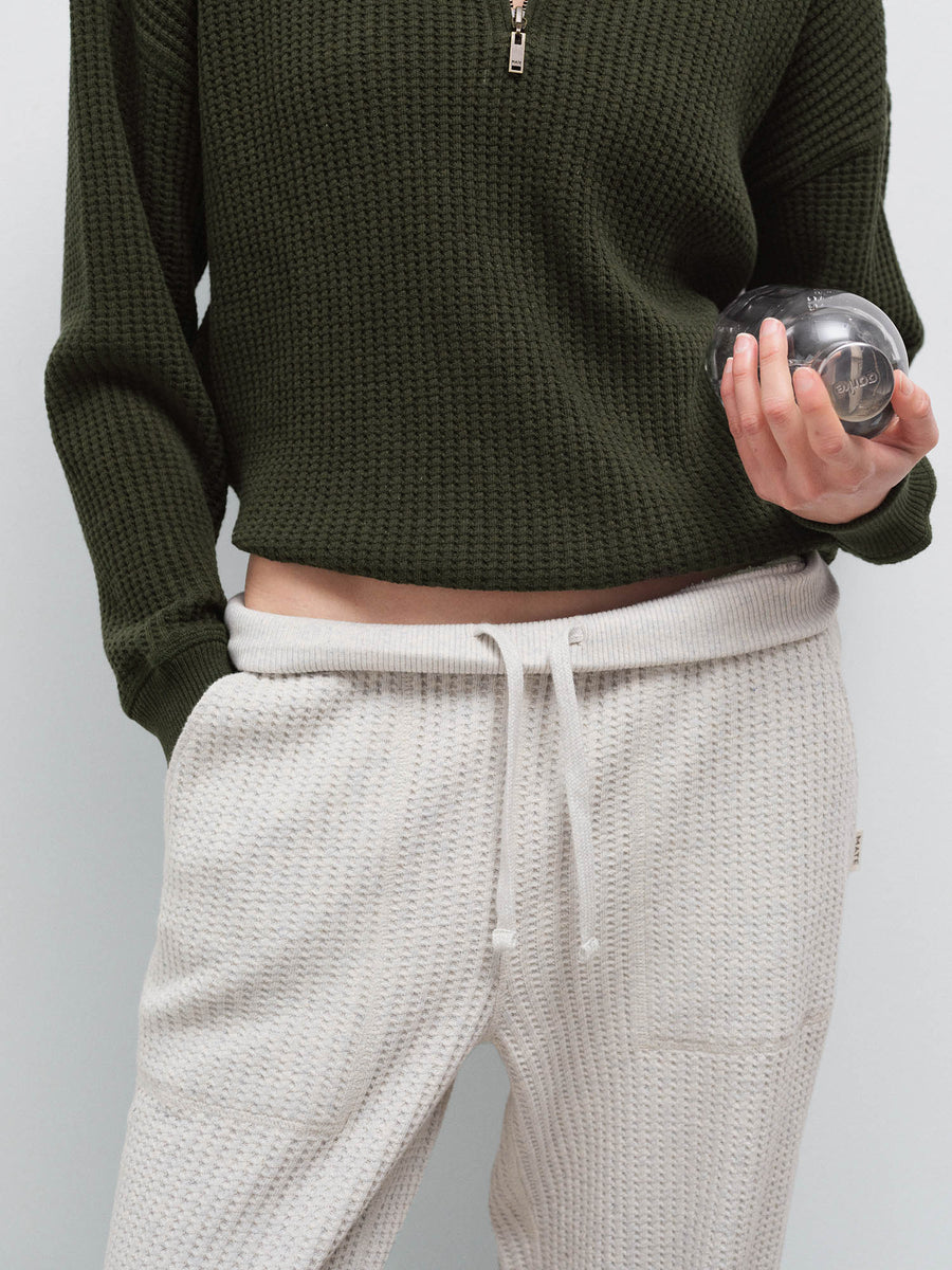Close-up of cream waffle-knit sweatpants with olive half-zip sweater hem, holding a small object in studio