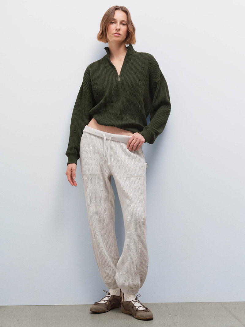 Woman wearing olive waffle-knit half-zip sweatshirt and cream sweatpants, full-length front view in studio