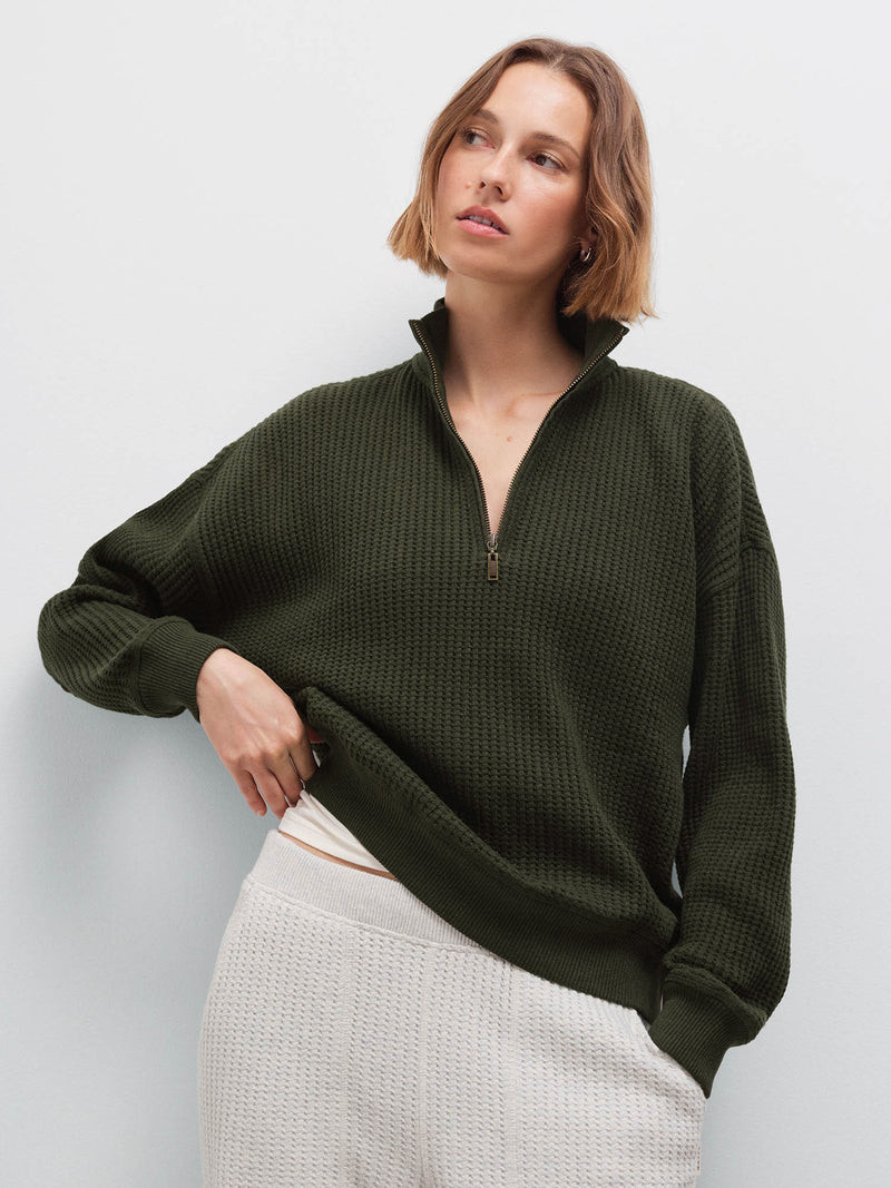 Woman wearing olive waffle-knit collared half-zip sweatshirt with cream sweatpants, front view in studio