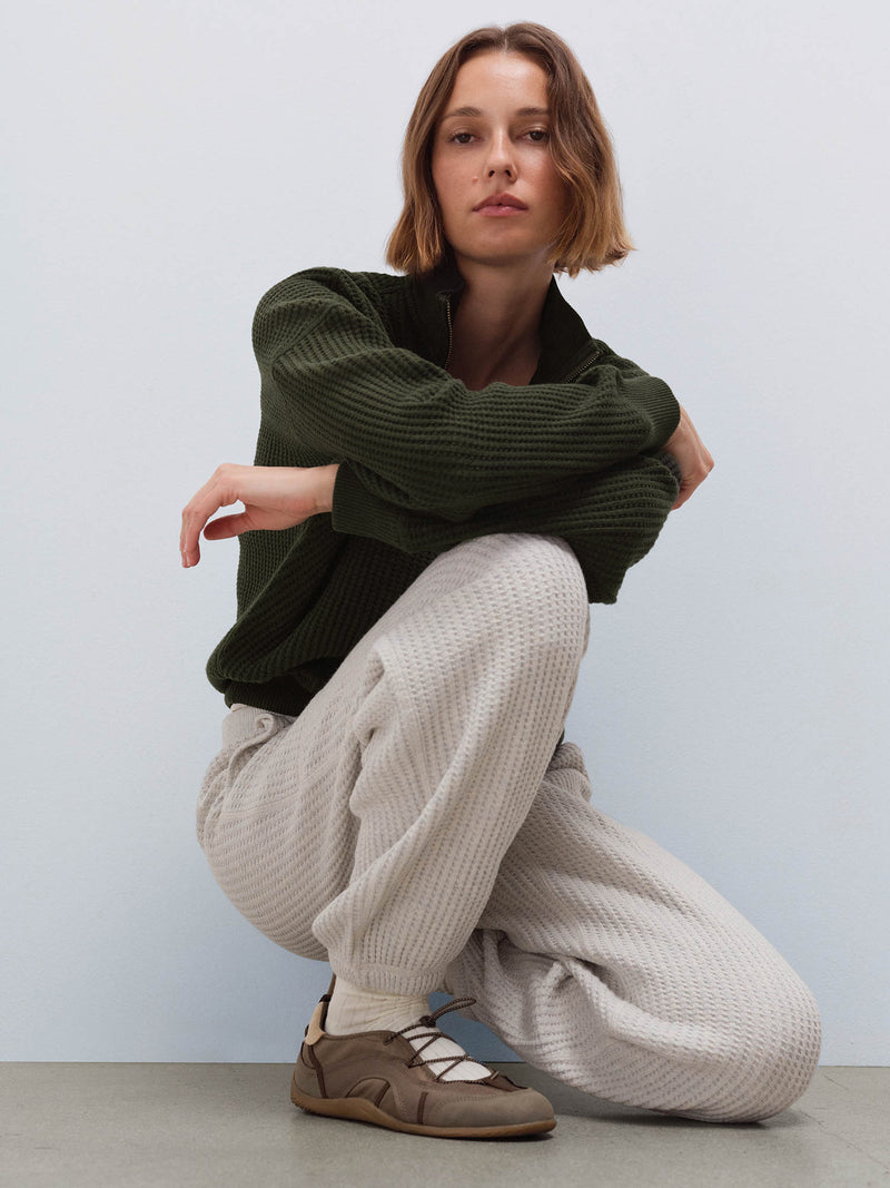 Woman wearing olive waffle-knit half-zip sweatshirt and cream sweatpants, crouching pose in studio