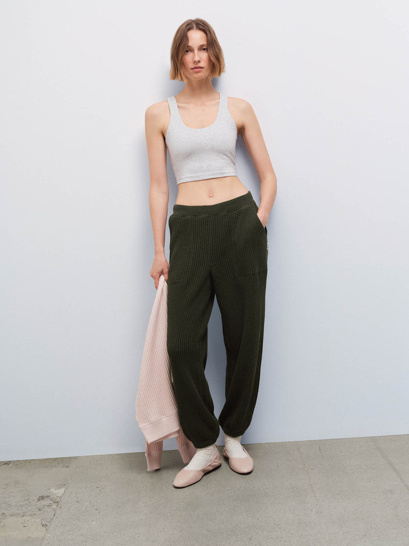 Woman wearing a heather gray tank and olive green waffle-knit jogger sweatpants, holding a pink cardigan, studio