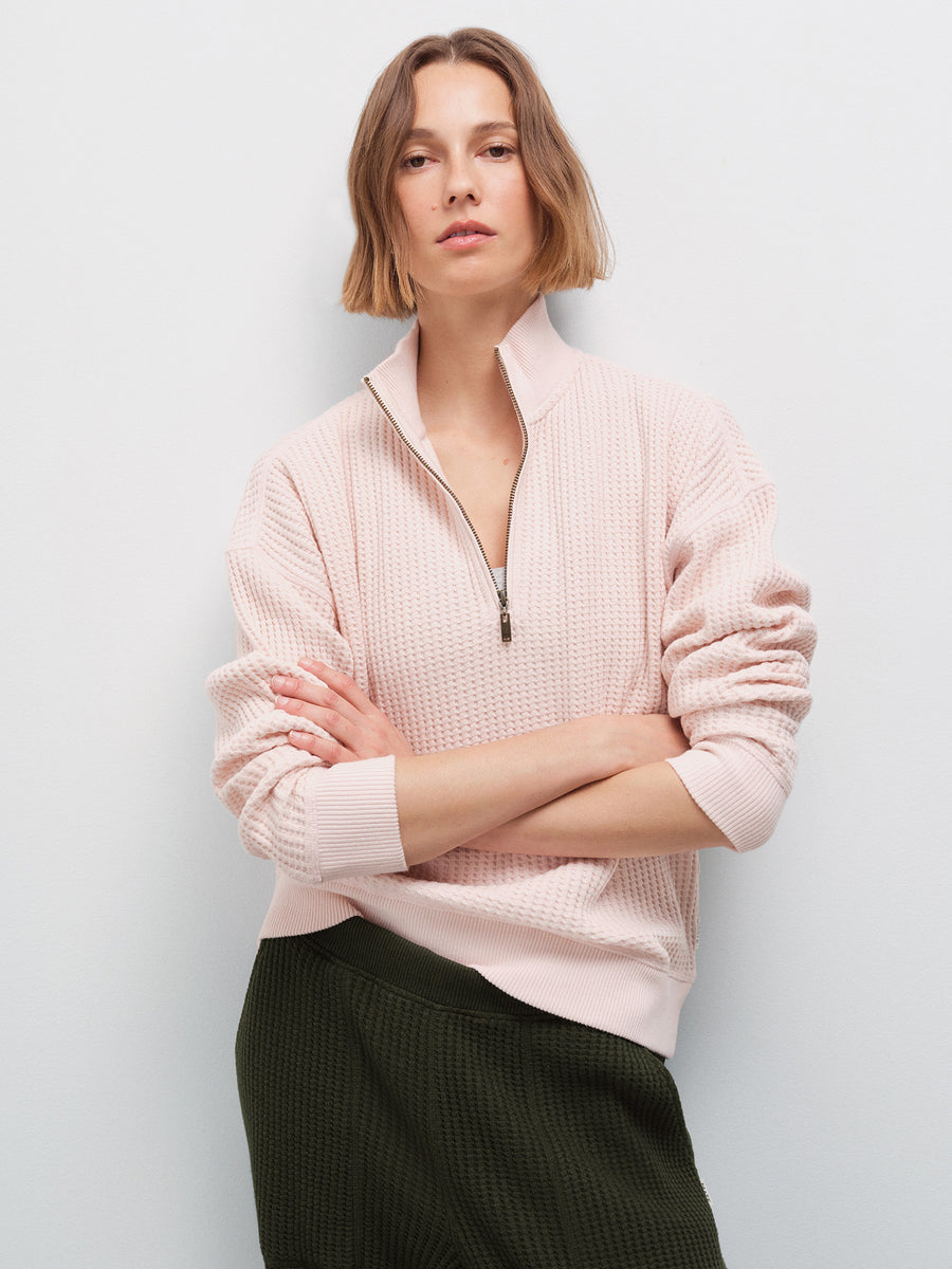 Woman with arms crossed wearing a pale pink waffle-knit quarter-zip pullover and olive green pants