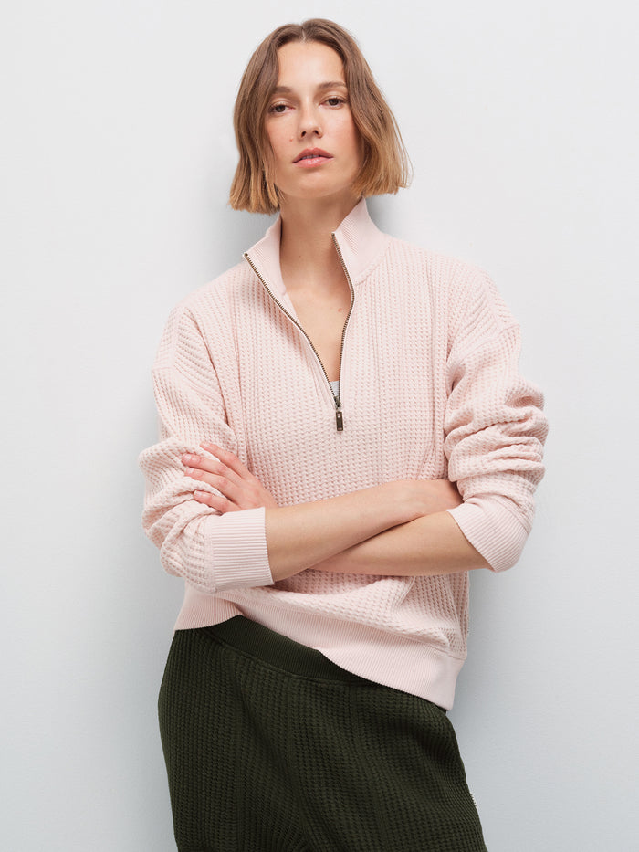 Woman with arms crossed wearing a pale pink waffle-knit quarter-zip pullover and olive green pants