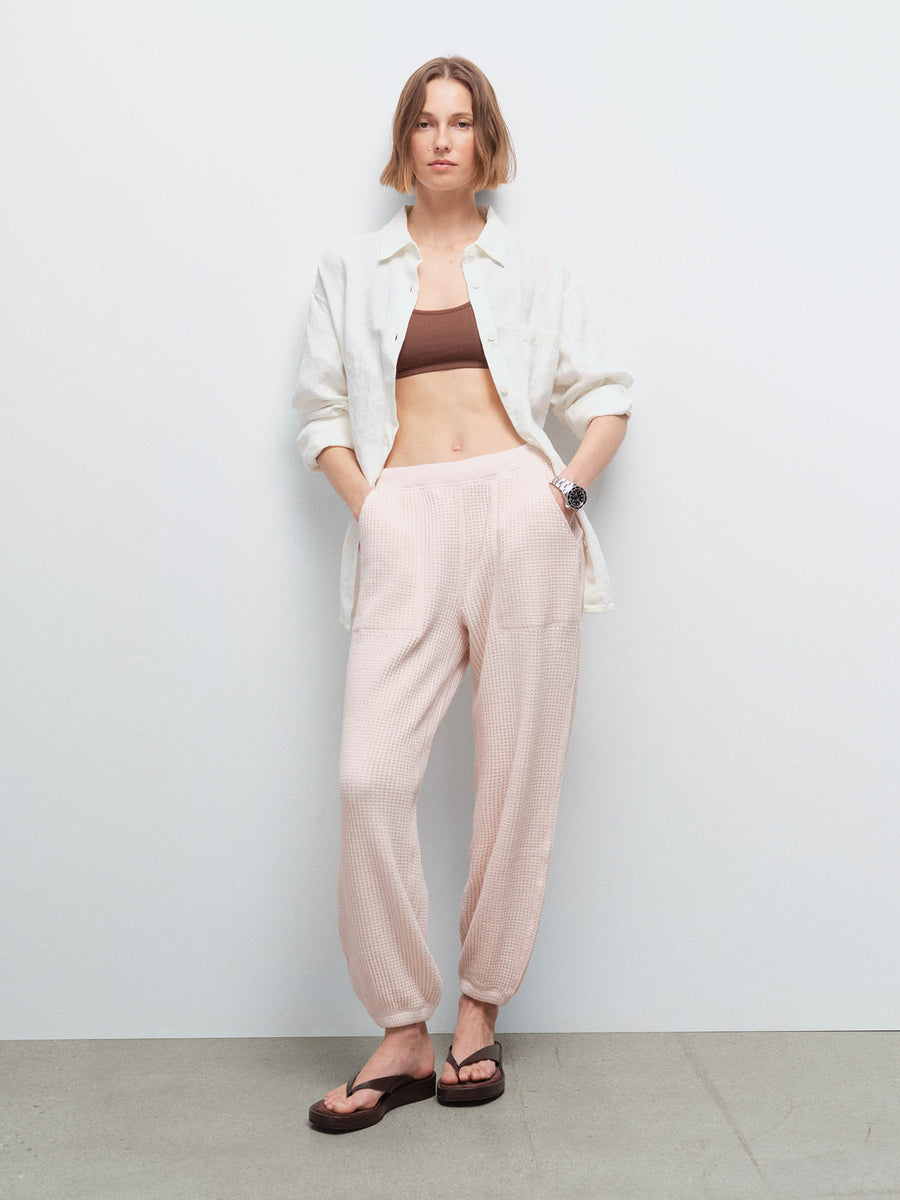Woman wearing an open cream shirt over a brown bralette and pale pink waffle jogger sweatpants with sandals, studio