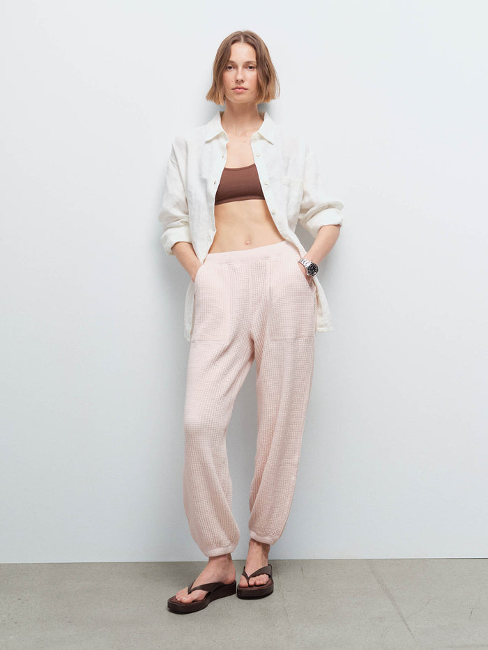 Woman wearing an open cream shirt over a brown bralette and pale pink waffle jogger sweatpants with sandals, studio