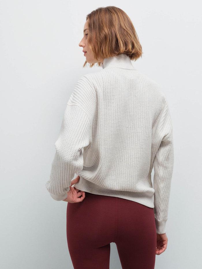 Woman standing wearing a cream waffle-knit quarter-zip pullover and burgundy leggings, back view in studio