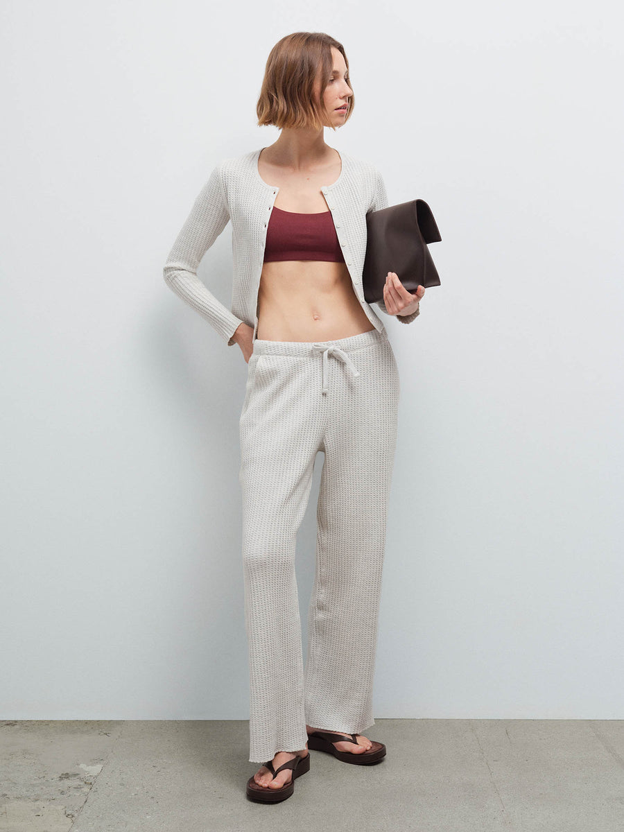 Woman wearing an open cream waffle-knit cardigan over a burgundy bralette with matching pants, holding a dark clutch
