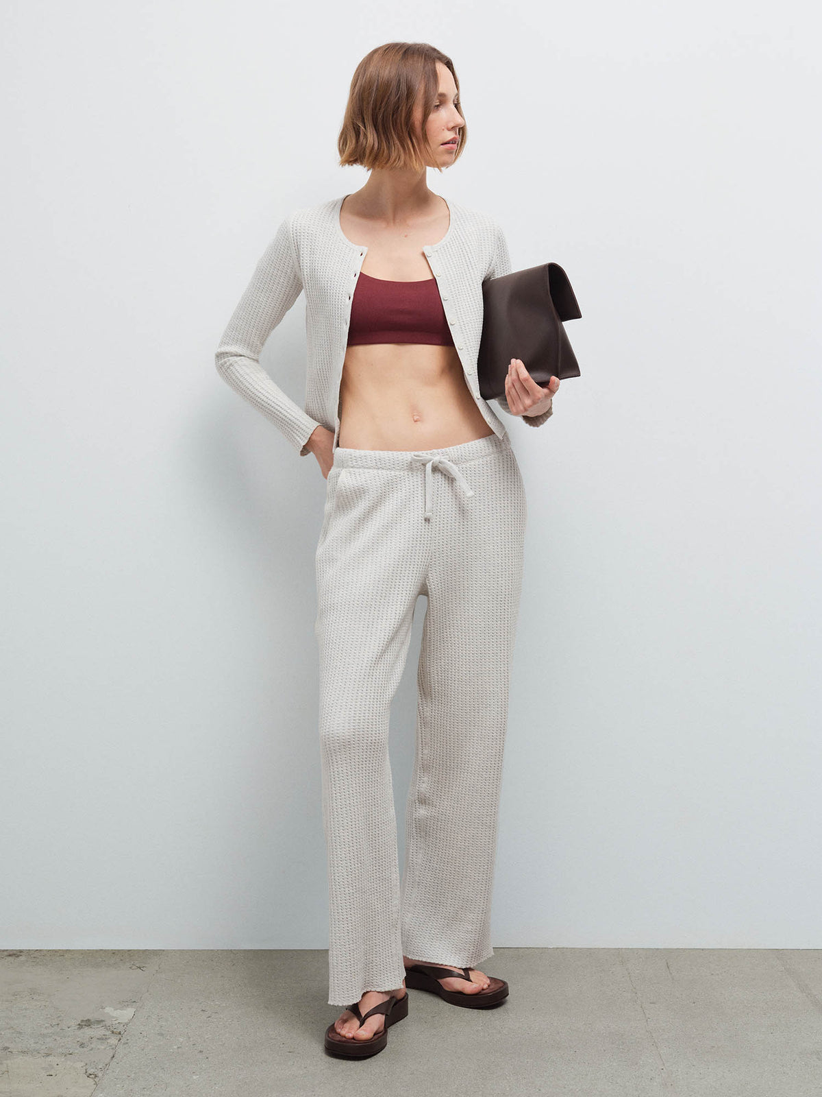 Woman wearing open cream waffle-knit cardigan over burgundy bra top with matching sweatpants, holding dark clutch, studio