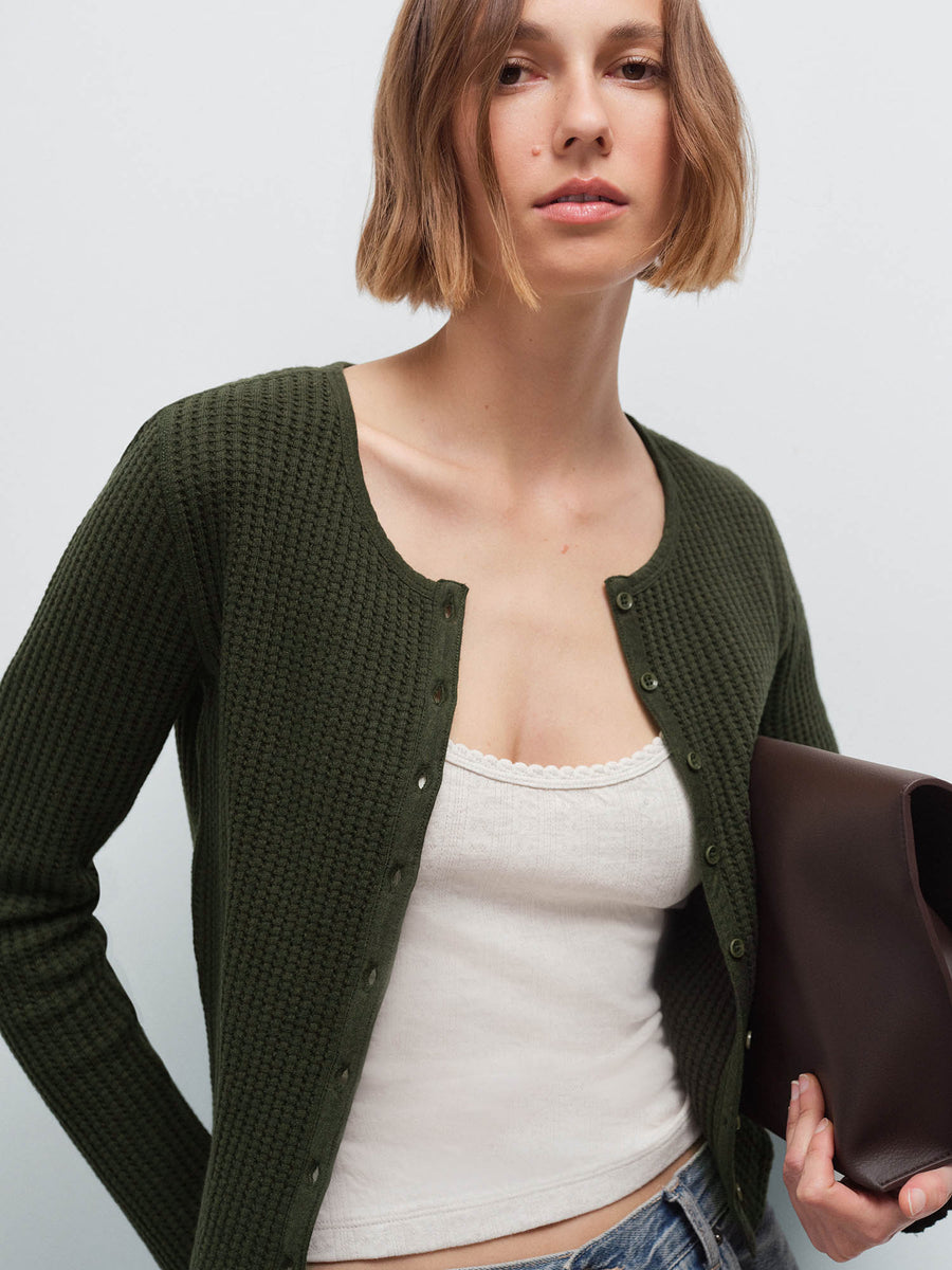 Close-up of a woman wearing an open olive green waffle-knit cardigan over a lace-trim cream tank, holding a brown clutch