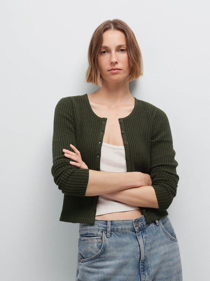 Close-up of a woman with arms crossed wearing an olive green waffle-knit cardigan over a cream top with denim jeans