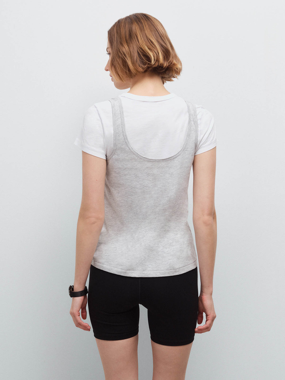 Woman standing wearing a heather grey tank layered over a white tee and black biker shorts, back view