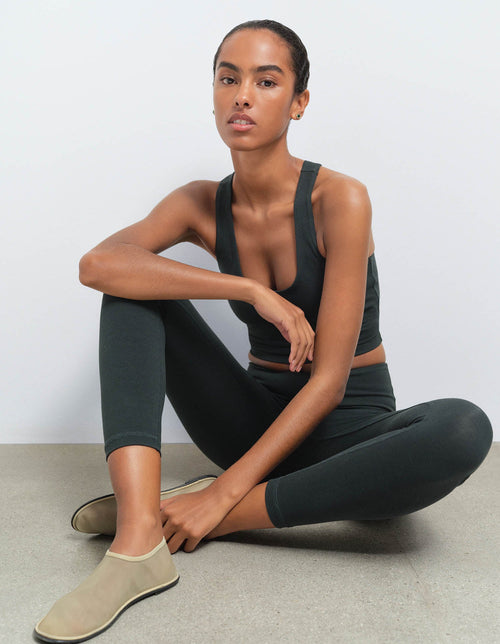 A woman sits cross-legged on a light gray floor against a white wall, wearing a dark olive green sports bra and matching leggings with beige slip-on shoes.