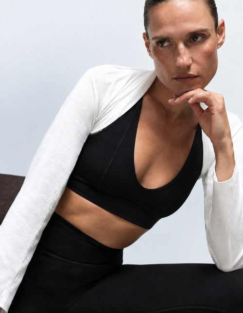 A woman wearing a black sports bra and black leggings with a white long-sleeve cardigan draped off her shoulders, posed against a light gray studio background.