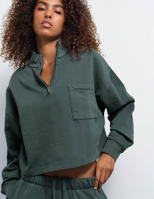 A woman wearing a dark green cropped quarter-zip sweatshirt with a chest pocket, paired with matching sweatpants, against a plain light gray background.