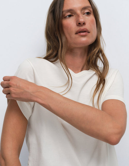 A woman wearing a plain off-white short-sleeve V-neck t-shirt, shown from the chest up against a light gray studio background.