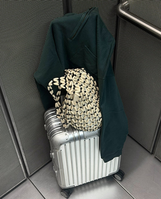 Dark green organic sweatshirt and woven bag on a silver suitcase in an elevator – MATE the Label